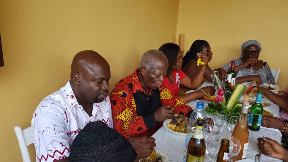 MBOGBE MBOGBE en boubou à motifs, partageant un repas convivial en famille