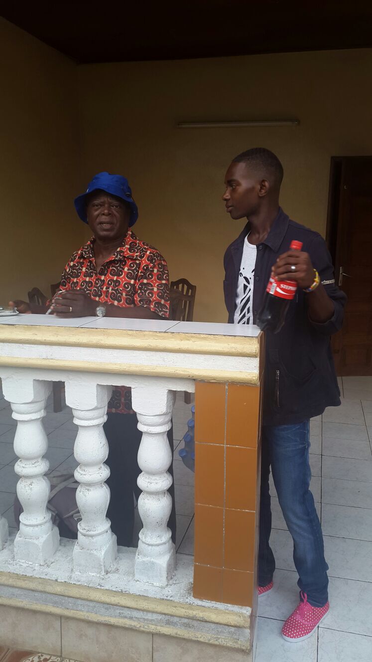 MBOGBE MBOGBE au chapeau bleu discutant avec un jeune sur une terrasse